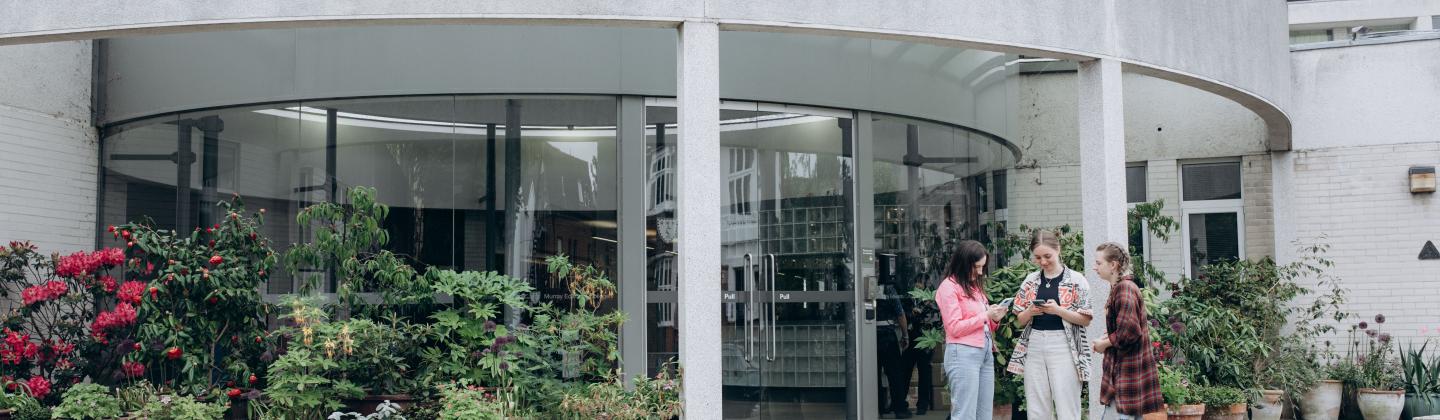 Steps lead up to a glass entrance to the College. At the top of the steps is a display of lush plants in large pots.