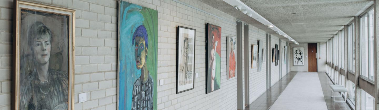Light-filled corridor with exposed brick wall on one side and large windows on the other. Artworks hung all along wall.
