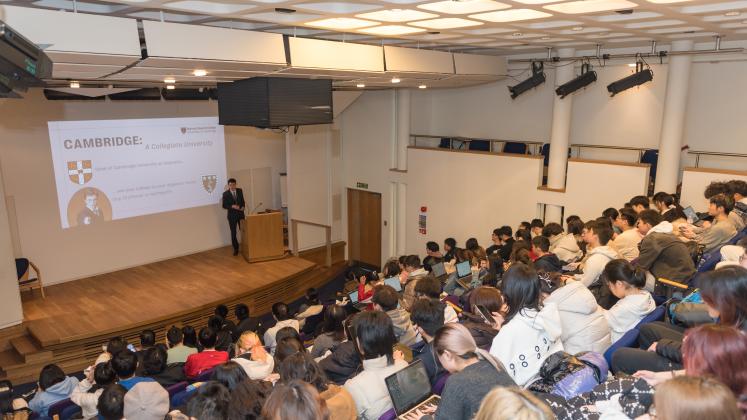 Lecture Theatre filled with students. From the stage a staff member gives a presentation about the college and university.