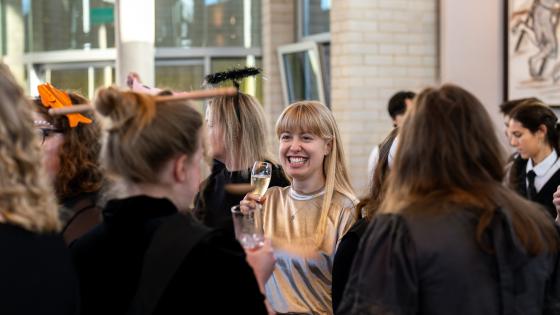 A woman standing with a glass of prosecco smiling. Others around her with waiters in the background.