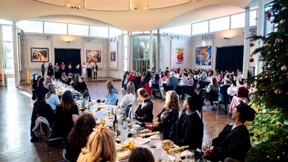 A long dining table with guests seated looking at a choir singing in the background. A Christmas tree in the foreground
