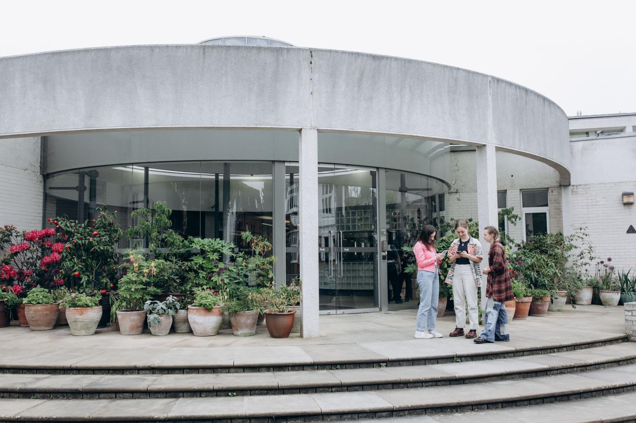 Steps lead up to a glass entrance to the College. At the top of the steps is a display of lush plants in large pots.