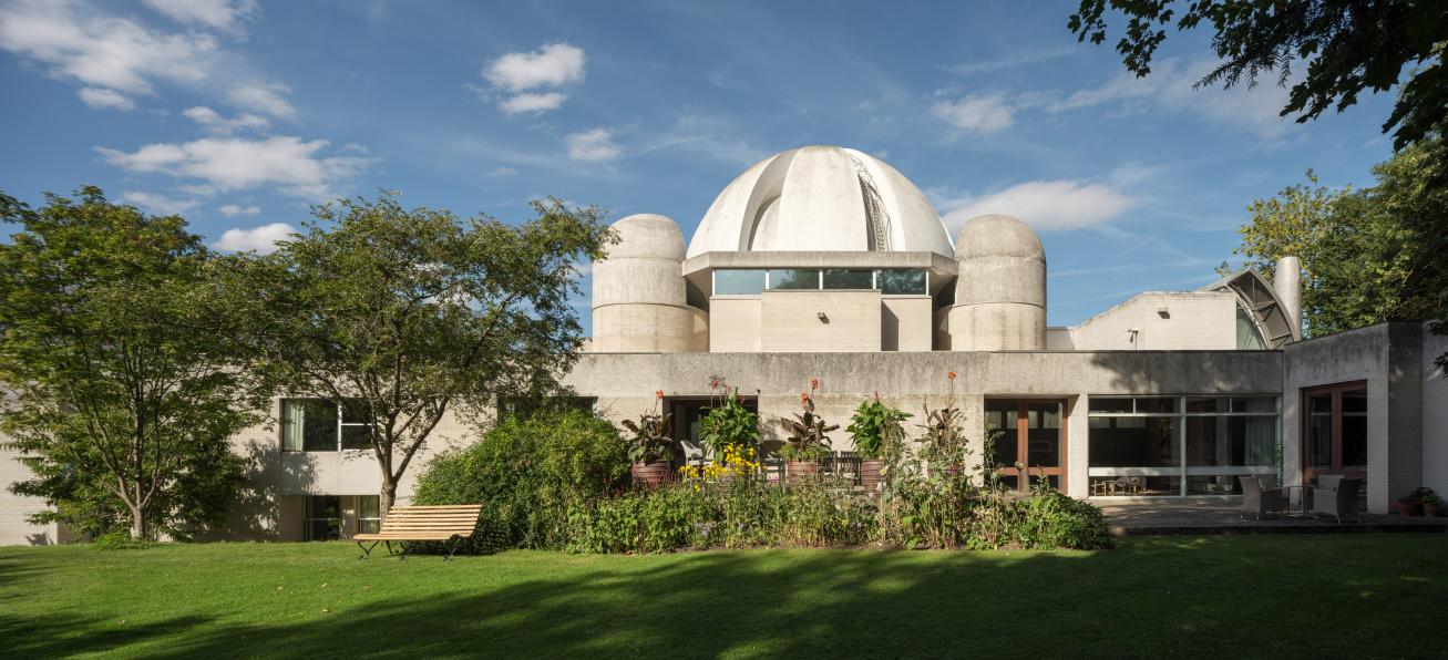 In the foreground, a grassed area. Behind, a patio with windows into the Fellows Drawing Room. Above, the a concrete dome of the dining hall