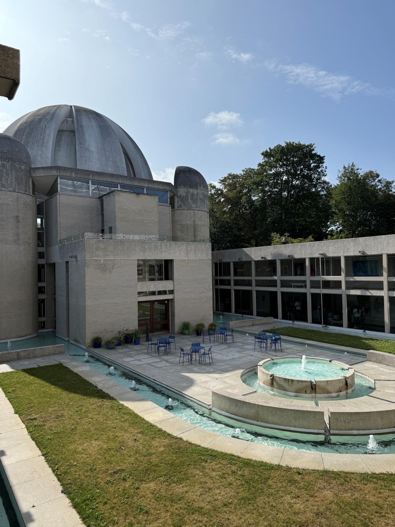 A building with a large, concrete dome as a roof. Below, a large water fountain with seating in the middle. To the side, a two floors of corridors with glass wall toward the fountain..